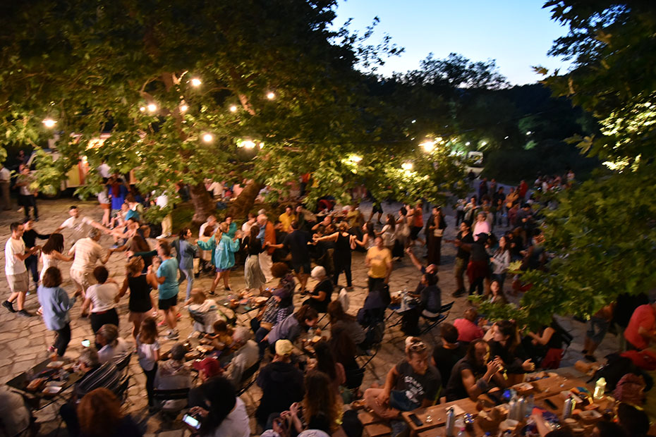 Μια ομάδα ανθρώπων που χορεύουν σε κύκλο σε μια πλατεία με μεγάλα δέντρα και λαμπερά φώτα 