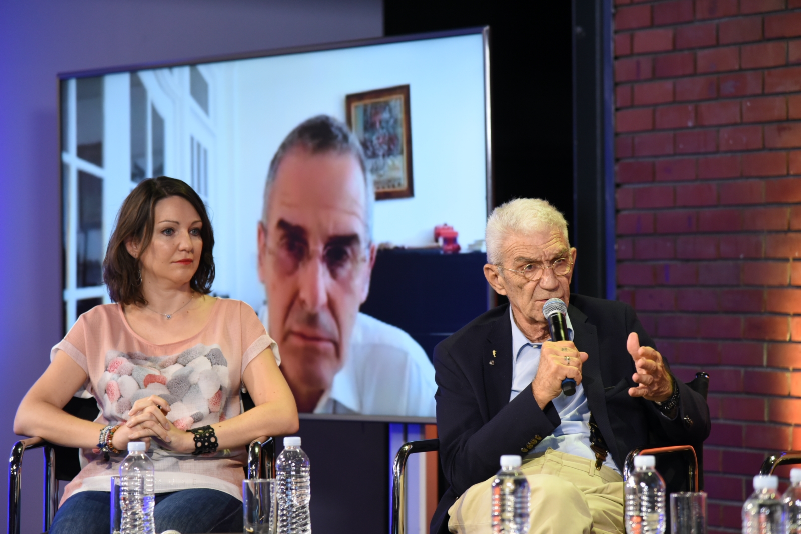 Δύο ομιλητές καθισμένοι σε καρέκλες και μια οθόνη που δείχνει ένα συμμετέχοντα στις Συζητήσεις των Διαλόγων ΙΣΝ.