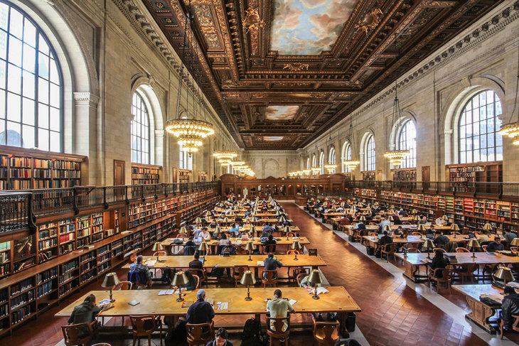 Α reading room of the New York Public Library filled with people studying at tables surrounded by countless books.