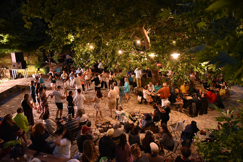 Μια ομάδα ανθρώπων που χορεύουν σε κύκλο σε μια πλατεία με μεγάλα δέντρα και λαμπερά φώτα 