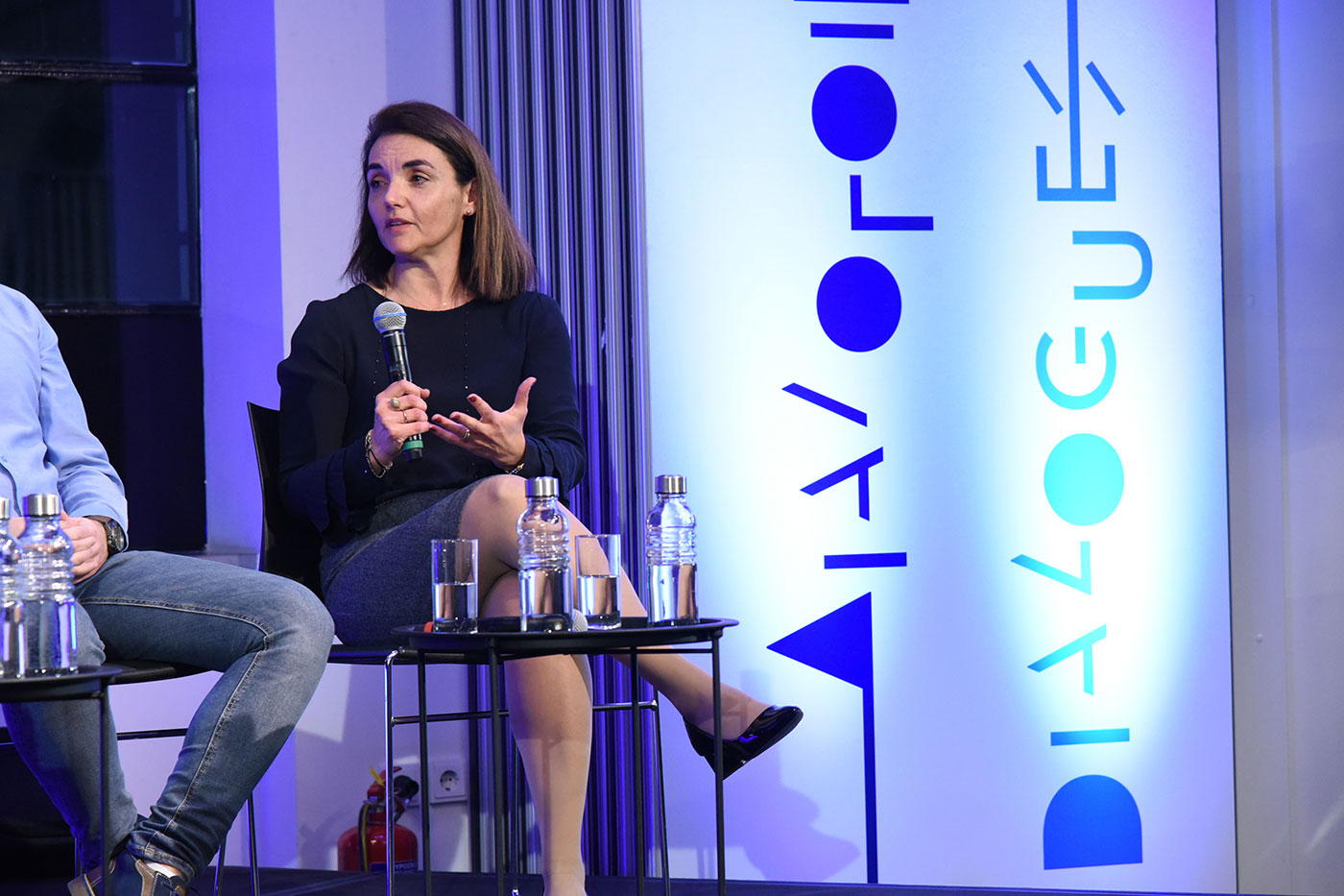 Μια καλεσμένη μιλάει στη συζήτηση των Διαλόγων ΙΣΝ μπροστά σε μια μεγάλη οθόνη με μπλε γράμματα