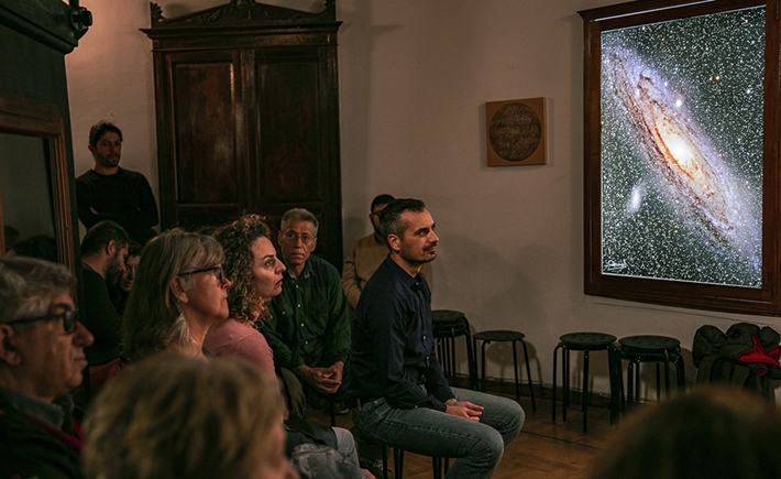 Άνθρωποι καθιστοί στις καρέκλες τους και ένα κάδρο με έναν πλανήτη πίσω τους 