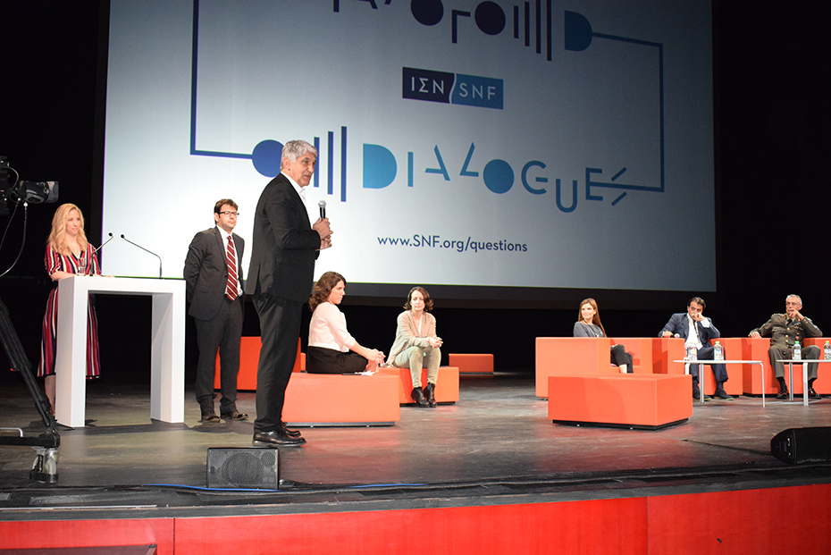 Ομιλητής στη σκηνή με πολλούς συμμετέχοντες στο πίσω μέρος στη συζήτηση των Διαλόγων ΙΣΝ.