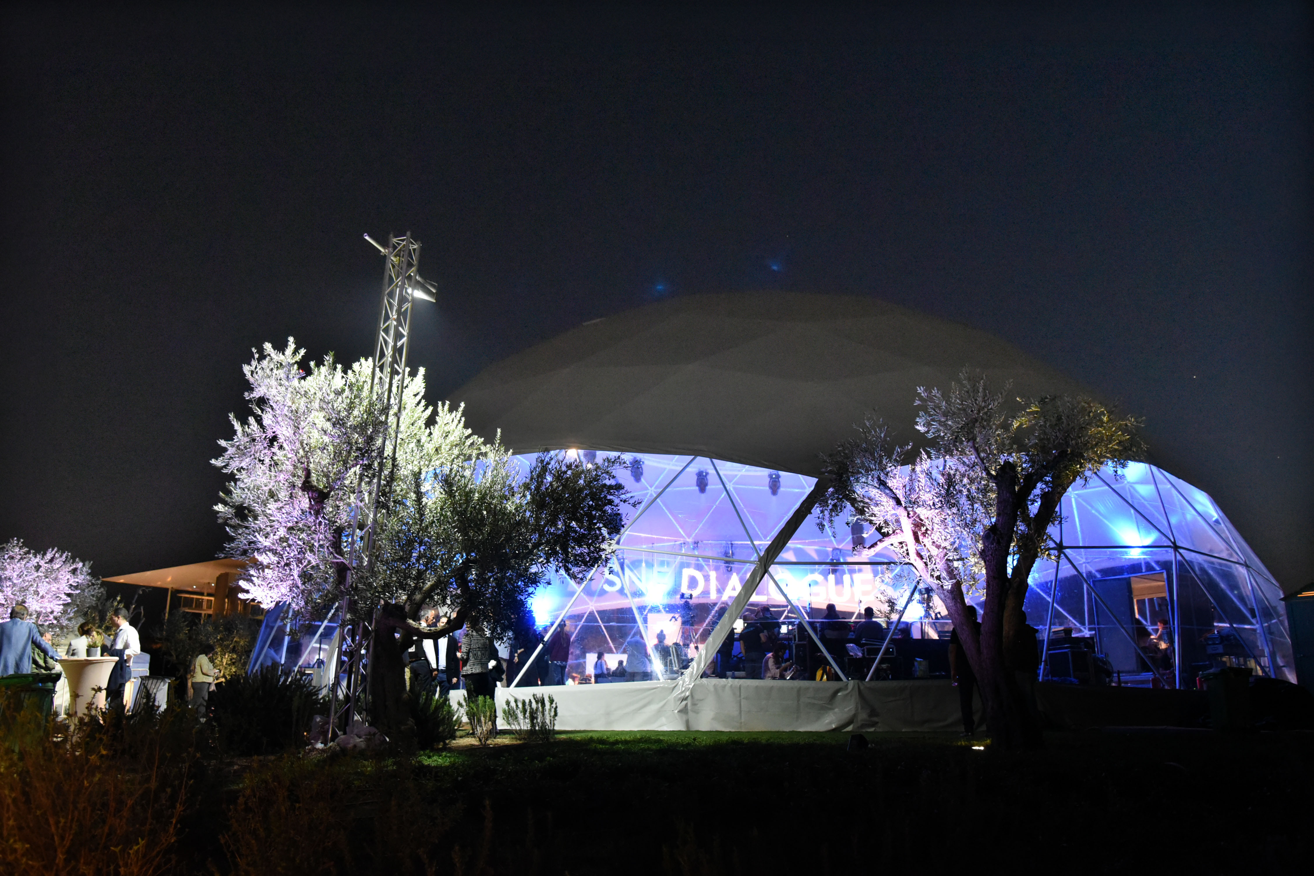 Άνθρωποι στέκονται γύρω και μέσα σε μια θολωτή σκηνή όπου πραγματοποιείται η συζήτηση των Διαλόγων ΙΣΝ.