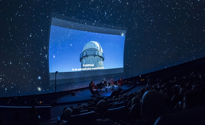 Καλεσμένοι ομιλητές με μια μεγάλη οθόνη πίσω τους με εικαστικά από το διάστημα και το κοινό που τους ακούει στη συζήτηση των Διαλόγων ΙΣΝ 