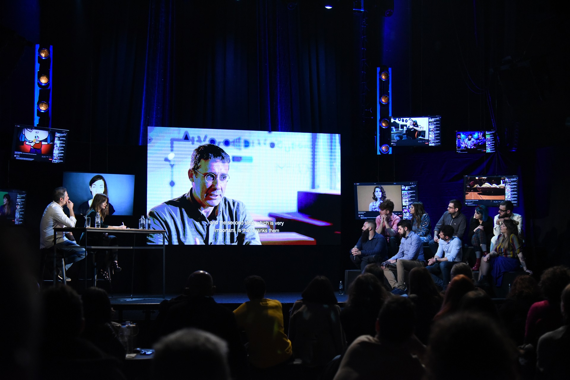 Κοινό παρακολουθεί μια παρουσίαση-βίντεο στη σκηνή στη συζήτηση Διαλόγων ΙΣΝ.