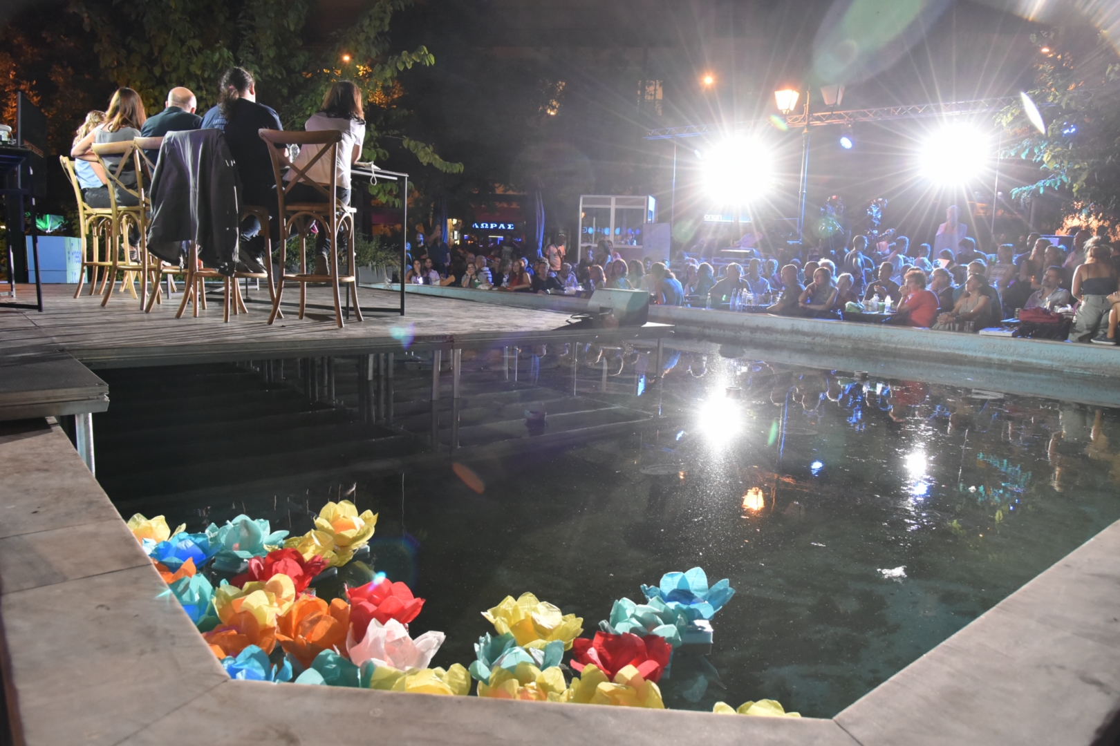  Close view of a pool with floating paper flowers, speakers on stage and audience in the background.