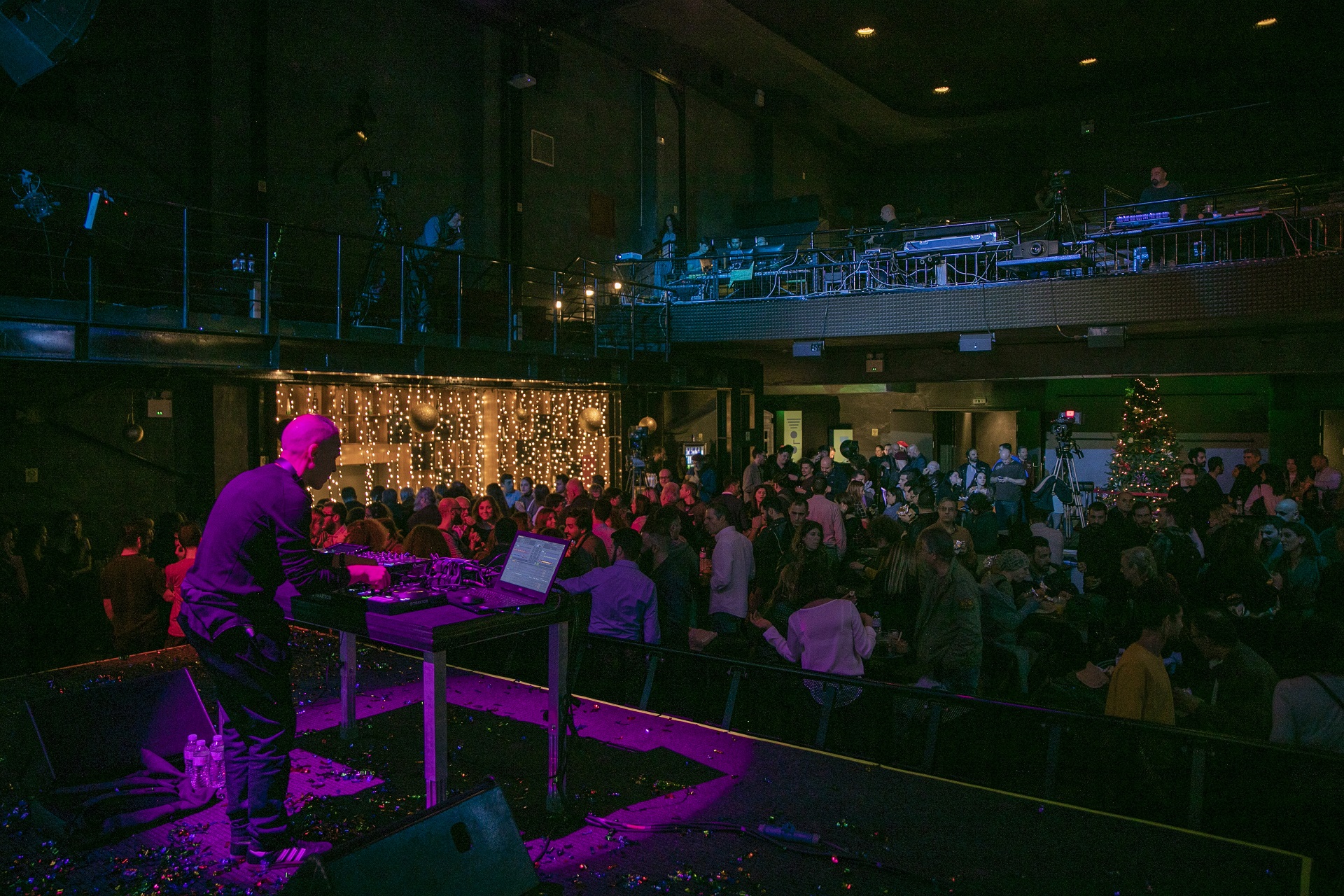 Ντι Τζέι στη σκηνή χειρίζεται την κονσόλα και κάτω από τη σκηνή μεγάλο κοινό σε έναν χριστουγεννιάτικα διακοσμημένο χώρο.