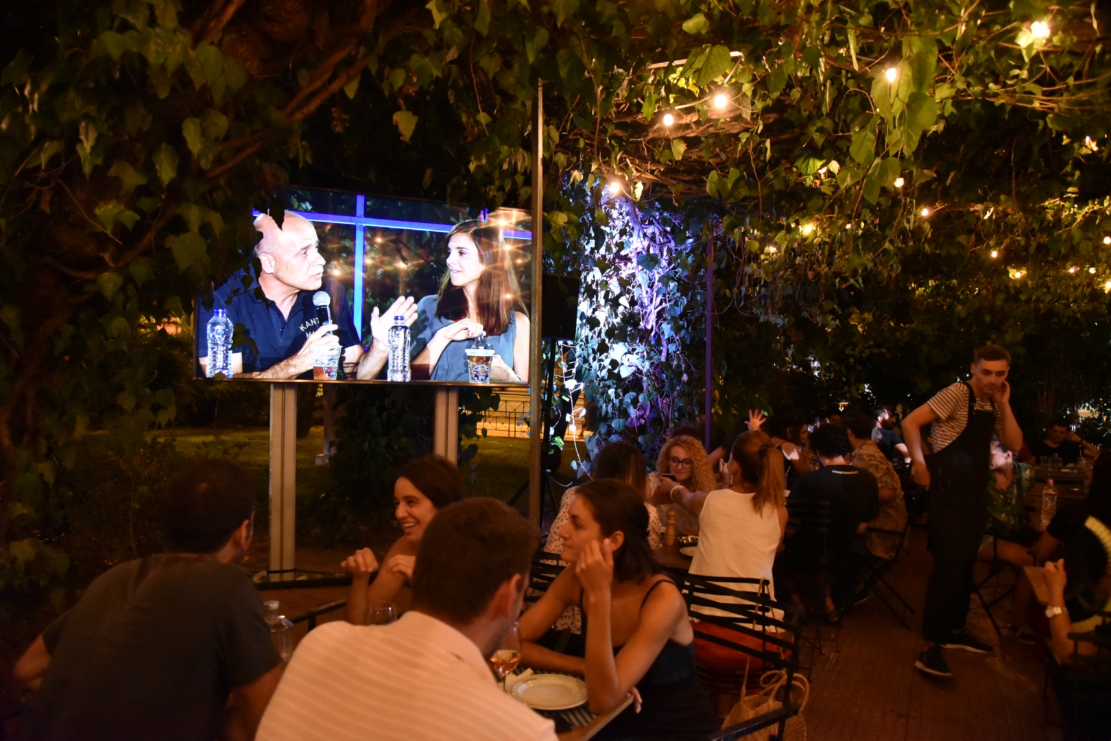 People sitting at tables in an outdoor area with a TV displaying the SNF Dialogues.