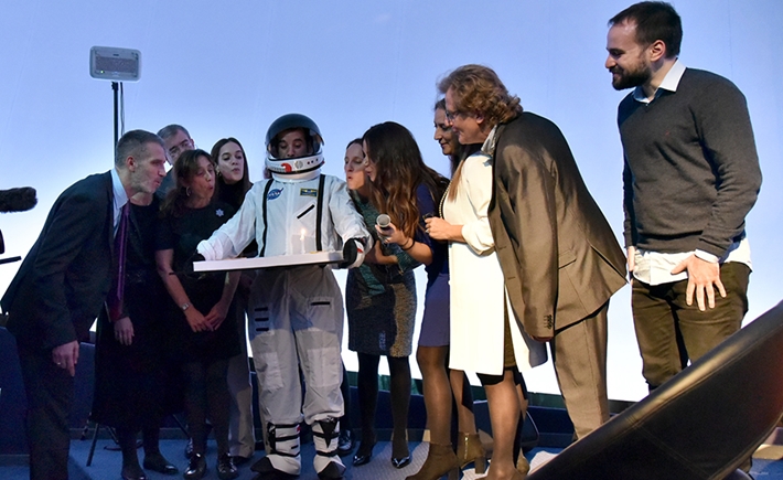 Ένας αστροναύτης που κρατάει μια τούρτα περιτριγυρισμένος από ανθρώπους που σβήνουν το κερί