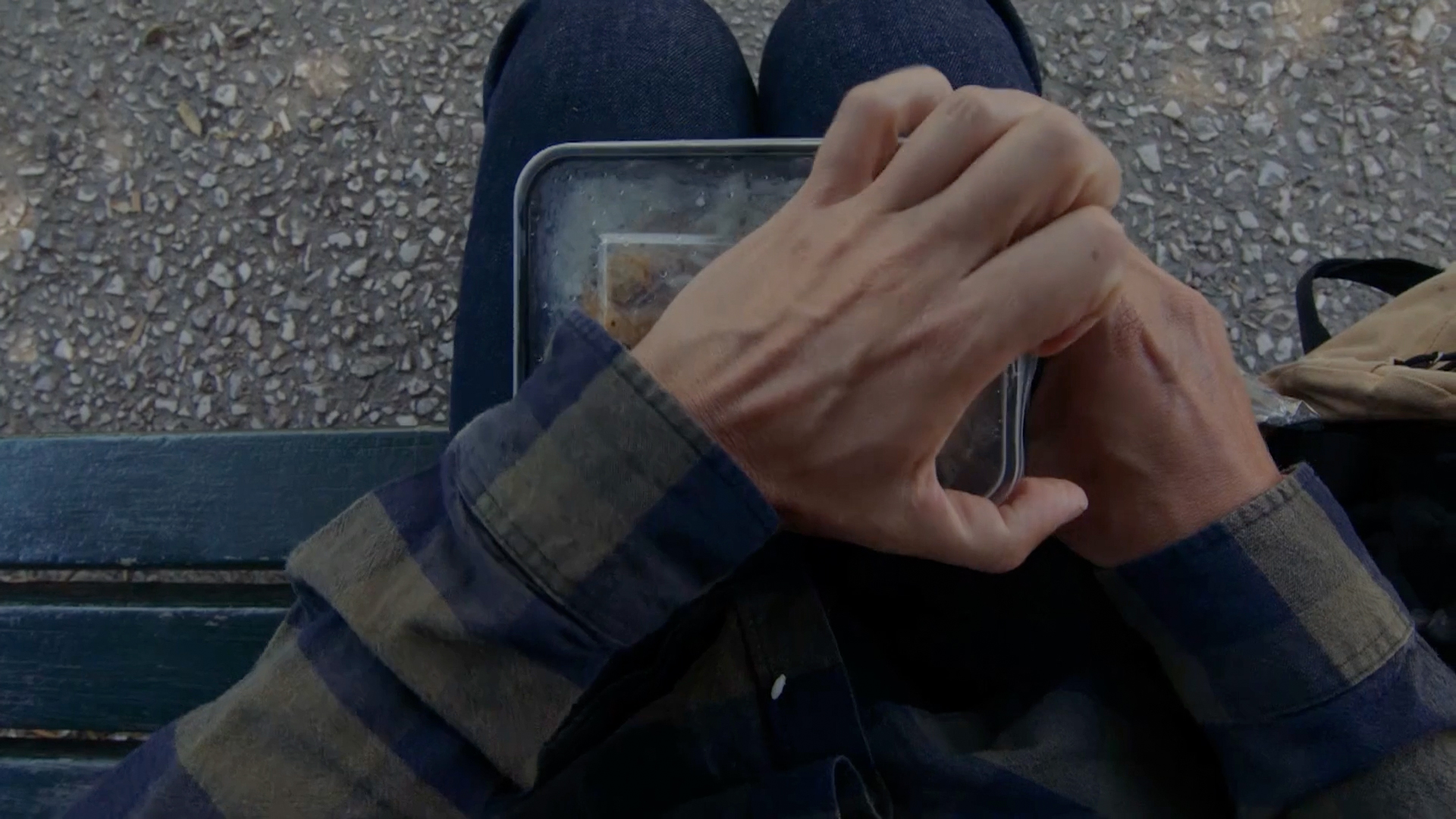 Ένας άνδρας που κάθεται σε ένα παγκάκι και ανοίγει ένα μικρό τάπερ με φαγητό 