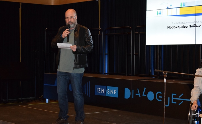 Ο Ανδρέας Δρακόπουλος, Πρόεδρος του ΙΣΝ, στέκεται στη σκηνή κρατώντας το μικρόφωνο και μιλά στο κοινό στη συζήτηση των Διαλόγων ΙΣΝ.