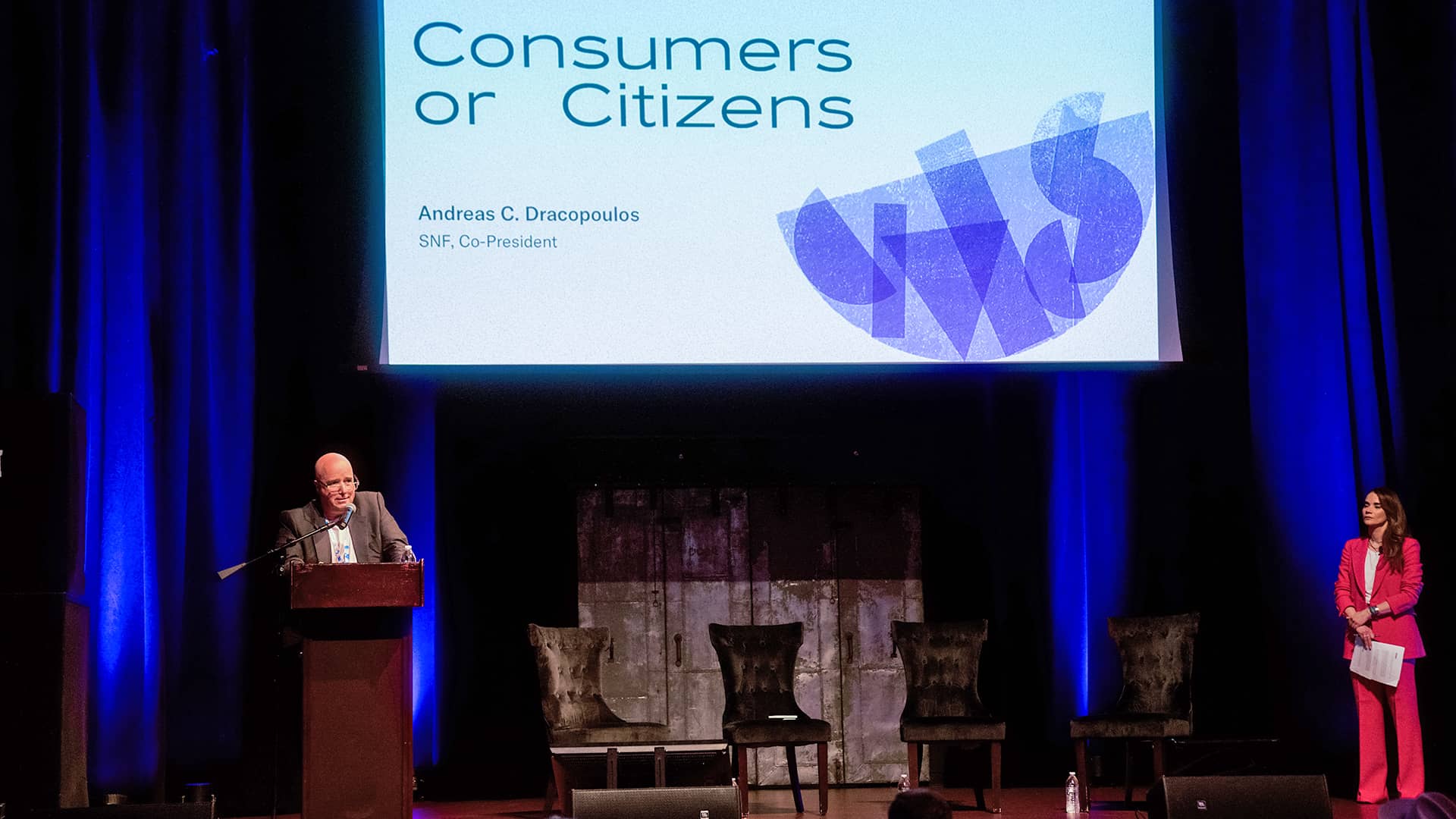 Ο Ανδρέας Δρακόπουλος εκφωνεί ομιλία και η Άννα-Κύνθια Μπουσδούκου στέκεται στη σκηνή. 