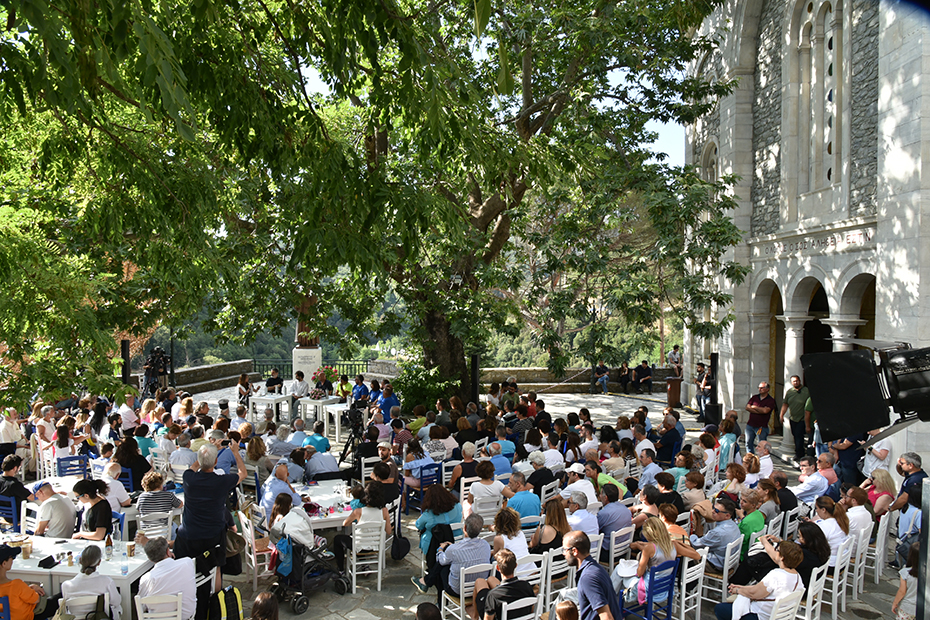 Ένα μεγάλο πλήθος παρευρισκομένων που κάθεται έξω από μια εκκλησία στο φως της ημέρας περιτριγυρισμένο από ένα μεγάλο δέντρο