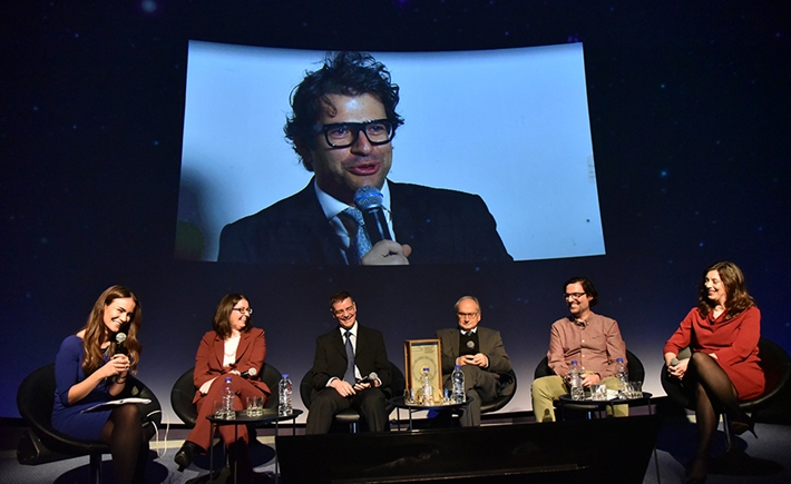 Έξι καλεσμένοι ομιλητές πάνω στην σκηνή και ένα κοντινό πλάνο ενός άνδρα που μιλάει σε μια μεγάλη οθόνη πίσω τους στη συζήτηση των Διαλόγων ΙΣΝ
