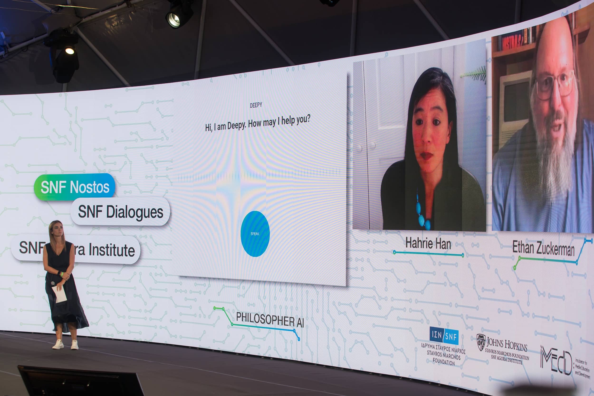 Anna Kynthia Bousdoukou on stage with a big screen behind her showing two speakers at the SNF Dialogues event.